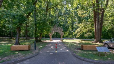 Shaded asphalt walking path winding through a densely wooded section of Overton Park in Midtown Memphis, featuring a decorative stone archway entry and wooden benches