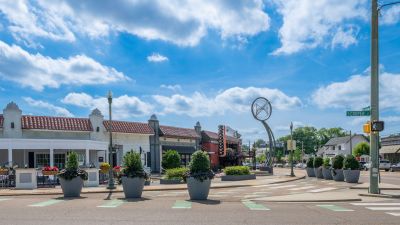Street view of Overton Square at the corner of South Cooper Street in Midtown Memphis, showcasing retail storefronts with red Spanish tile roofs, public art sculptures, and sidewalk decorative planters.