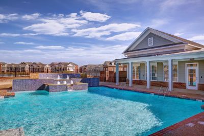 Resort-style swimming pool with fountain features and clubhouse at a Memphis apartments community, surrounded by residential homes under a blue sky