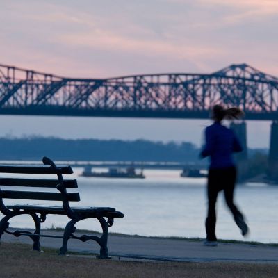 Person jogging along the Mississippi River at dusk near Downtown Memphis, with a historic bridge silhouetted against a pink and blue sunset sky