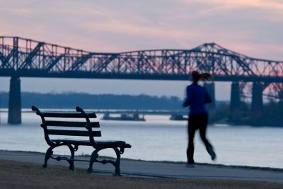 Person jogging along the Mississippi River at dusk near Downtown Memphis, with a historic bridge silhouetted against a pink and blue sunset sky