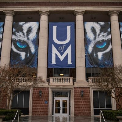 Front facade of a University of Memphis campus building displaying large blue and white tiger mascot banners and the U of M logo