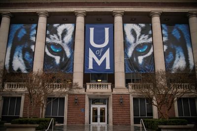 Front facade of a University of Memphis campus building displaying large blue and white tiger mascot banners and the U of M logo