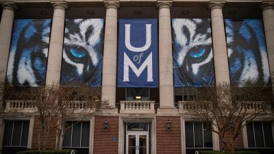 Front facade of a University of Memphis campus building displaying large blue and white tiger mascot banners and the U of M logo