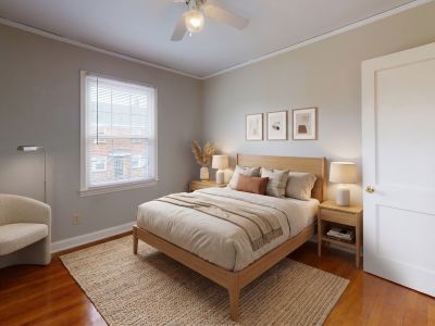 Bright staged bedroom showing warm hardwood flooring, neutral-colored linens, a rounded accent chair, and large windows at Audubon Downs apartments in Memphis.