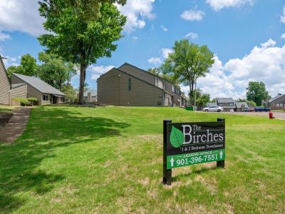 A green lawn showcasing a leasing office sign for The Birches in front of multi-level townhome buildings.