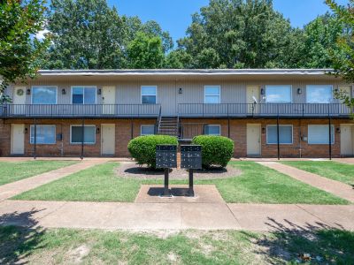 Symmetric front view of a two-story apartment building exterior with a brick lower level, beige upper siding, a continuous second-floor balcony with iron railings, centralized outdoor mailboxes, and manicured green hedges.