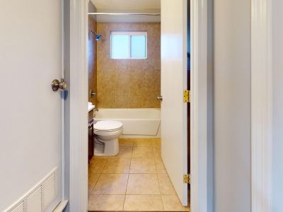 Straight-on view of a bathroom interior looking through the doorway, featuring tan floor tiles, a white toilet, and a standard bathtub with a matching tan-tiled wall surround and an overhead frosted window.