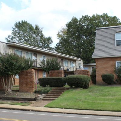 Street-side perspective of several multi-family apartment buildings showing a mix of brick facings, mansard roof styling, a well-kept green lawn, and brick retaining walls with stone steps leading up the slope
