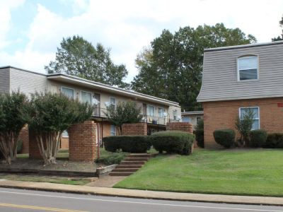 Street-side perspective of several multi-family apartment buildings showing a mix of brick facings, mansard roof styling, a well-kept green lawn, and brick retaining walls with stone steps leading up the slope