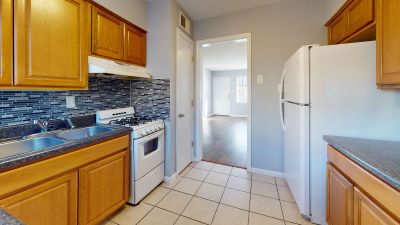 Corner view of an apartment kitchen featuring honey-oak wood cabinetry, a multi-colored mosaic tile backsplash, a white refrigerator, a white gas stove, and an open view looking into a living room with wood-style plank flooring
