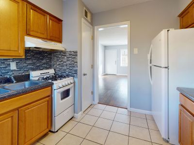 Corner view of an apartment kitchen featuring honey-oak wood cabinetry, a multi-colored mosaic tile backsplash, a white refrigerator, a white gas stove, and an open view looking into a living room with wood-style plank flooring