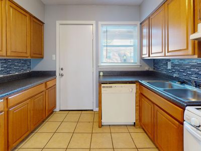 Galley-style view of the kitchen showcasing wooden cabinetry and dark countertops on both sides, a white built-in dishwasher, a double basin stainless steel sink, and a plain white interior door.