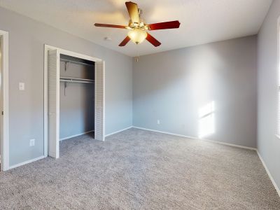 Bright, carpeted bedroom interior with light gray walls, a central multi-blade ceiling fan, a window with mini-blinds, and an open closet featuring white bi-fold slatted doors.