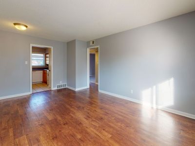 Unfurnished, open-concept living or dining space highlighting polished wood-look laminate floors, neutral gray walls, a simple ceiling light fixture, and an entryway leading back toward the kitchen tile floor.