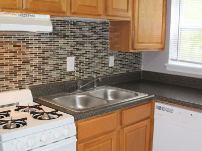 Close-up kitchen shot detailing the white gas cooking range, a white built-in dishwasher, a double stainless steel sink, and a modern mosaic glass tile backsplash paired with honey-oak cabinets.