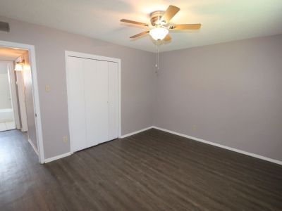 Unfurnished room highlighting dark wood-style flooring, light grey walls, a ceiling fan, white bi-fold closet doors, and a hallway view leading to a bathroom at Center Court Apartments in Collierville, TN.