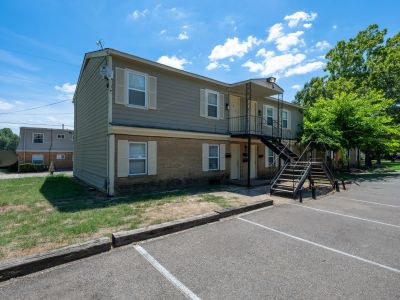 Exterior view of a two-story residential building featuring a brick lower level, grey siding upper level with decorative window shutters, an external black metal staircase, and an asphalt parking area with log bumper blocks at Center Court Apartments in Collierville, TN.