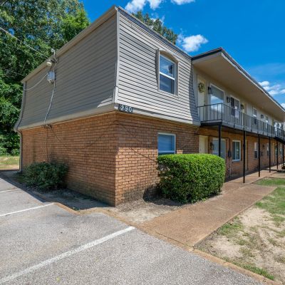 Corner profile of a multi-family building at building number 290, highlighting the brick exterior, mansard siding upper level, a concrete sidewalk with neat green hedges, and an adjacent parking lot at Center Court Apartments in Collierville, TN.