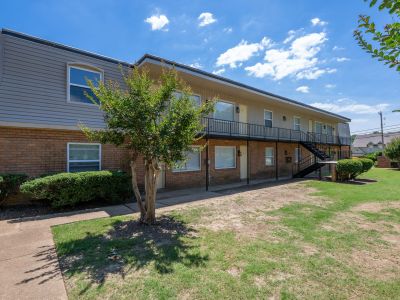 Ground-level perspective looking across a grassy courtyard lawn at a two-story brick and siding building with second-floor open walkways, featuring a young shade tree in the foreground at Center Court Apartments in Collierville, TN.