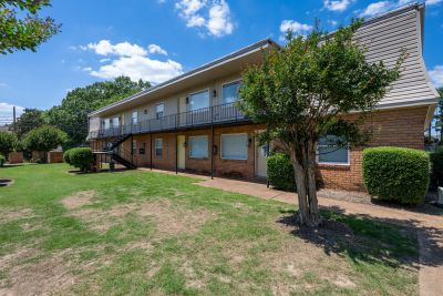 Courtyard view from an opposite angle showcasing a small tree, trimmed spherical bushes, and the front facade of a two-story apartment wing with long balconies under a bright blue sky at Center Court Apartments in Collierville, TN.