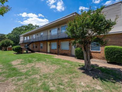 Courtyard view from an opposite angle showcasing a small tree, trimmed spherical bushes, and the front facade of a two-story apartment wing with long balconies under a bright blue sky at Center Court Apartments in Collierville, TN.