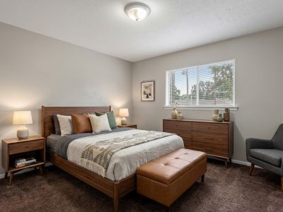 Staged primary bedroom showcasing mid-century modern wooden furniture, a leather storage bench, and deep brown carpeting at Cherry Creek apartments in Memphis.