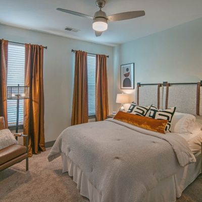 Designer apartment bedroom layout with copper-toned window drapes, a leather armchair, plush carpeting, and a ceiling fan at Madison@McLean Midtown apartments in Memphis.