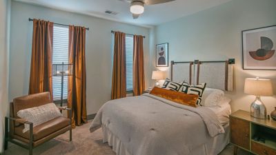 Designer apartment bedroom layout with copper-toned window drapes, a leather armchair, plush carpeting, and a ceiling fan at Madison@McLean Midtown apartments in Memphis.