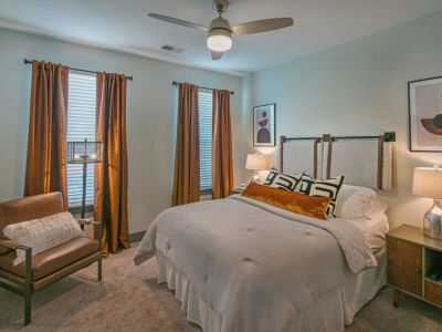 Designer apartment bedroom layout with copper-toned window drapes, a leather armchair, plush carpeting, and a ceiling fan at Madison@McLean Midtown apartments in Memphis.