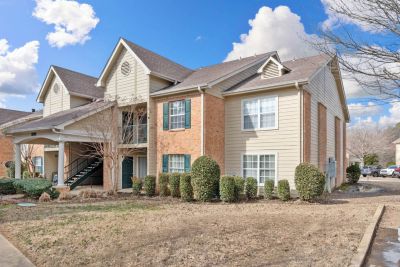 Exterior view of a multi-family apartment building with a light-toned brick and tan vinyl siding exterior, featuring dark green window shutters, white-trimmed multi-pane windows, and a covered walkway porch entrance under a bright blue sky.