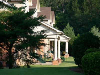 A scenic, angled perspective down a row of apartment building facades, showing ground-floor entry porches with classic white columns, decorative hanging flower baskets, and a deep green manicured lawn.