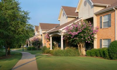 A smooth, winding concrete sidewalk paths through a wide green courtyard common area, passing by residential buildings accented with beautiful purple flowering trees.