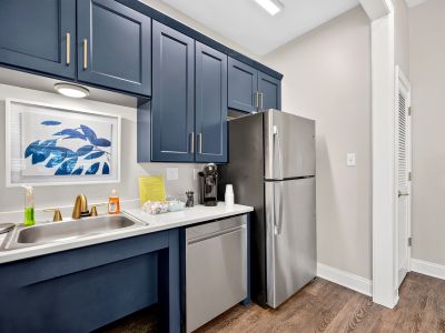 A clean, contemporary clubhouse kitchen or breakroom alcove designed with deep navy blue cabinets, brushed gold handles, stainless steel appliances, a coffee maker, and an abstract blue leaf art print.