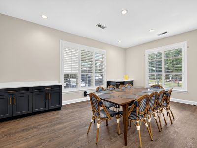 Spacious community room or meeting space featuring a long wooden conference table surrounded by woven blue and white chairs, dark cabinetry with white countertops, and plantation shutters over two windows at The Magnolias apartments in Hernando, MS.