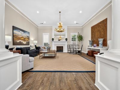 Grand clubhouse lounge viewed through architectural columns, showcasing high ceilings with an antler chandelier, a central fireplace, a large woven wall sculpture, plush dark sofas, and a large area rug at The Magnolias apartments in Hernando, MS.