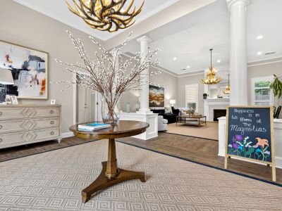 Clubhouse reception lobby showing a central round table with decorative willow branches, an antler chandelier, and a welcoming blackboard sign reading 'Happiness is in full bloom at The Magnolias' at The Magnolias apartments in Hernando, MS.