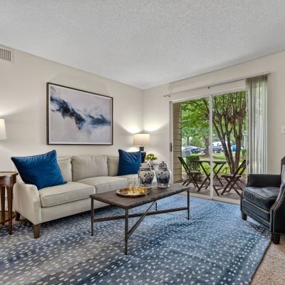 Comfortable apartment living room featuring a neutral sofa with deep blue accent pillows, a blue geometric area rug, a dark leather armchair, and a sliding glass door opening to a private patio at The Magnolias apartments in Hernando, MS.