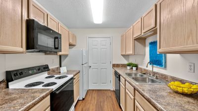 Galley-style apartment kitchen featuring light oak cabinetry, textured brown countertops, white appliances, a black over-the-range microwave, and wood-look laminate flooring at The Magnolias apartments in Hernando, MS.