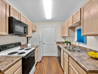 Galley-style apartment kitchen featuring light oak cabinetry, textured brown countertops, white appliances, a black over-the-range microwave, and wood-look laminate flooring at The Magnolias apartments in Hernando, MS.