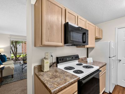 Close-up of a kitchen workspace detailing the light oak upper and lower cabinets, white range stove, black microwave, and an open layout peering directly into the carpeted living area at The Magnolias apartments in Hernando, MS.