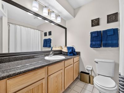 Clean apartment bathroom featuring an extended light oak vanity with a black granite-look countertop, a large framed mirror, a white toilet, and decorative navy blue towels at The Magnolias apartments in Hernando, MS.