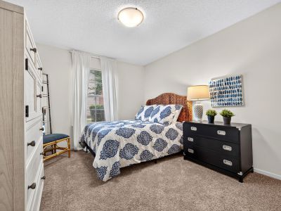 Furnished bedroom setup showing a wicker headboard, blue and white patterned bedding, a tall white armoire, and a wide window with long white curtains at The Magnolias apartments in Hernando, MS.