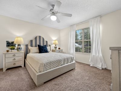 Bedroom showcasing an upholstered striped headboard, matching nightstands with blue and white porcelain lamps, a multi-blade ceiling fan, and long white window curtains at The Magnolias apartments in Hernando, MS.