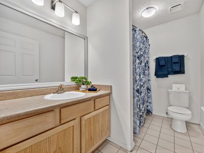 Clean apartment bathroom featuring a long light wood vanity with a framed mirror, a separate toilet alcove, a shower with a blue patterned curtain, and tan floor tiles at The Magnolias apartments in Hernando, MS.