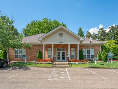 Front entrance of the brick leasing office building featuring architectural columns, landscaped flower beds, and a parking lot at The Magnolias apartments in Hernando, MS.