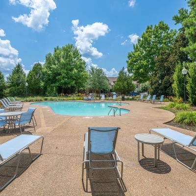 Resort-style community swimming pool with lounge chairs, a patio table set, and surrounding green trees under a blue sky at The Magnolias apartments in Hernando, MS.