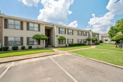 Two-story apartment building exterior featuring light vinyl siding, dark shutters, and manicured landscaping trees at The Ellington at Kirby apartments in Memphis.