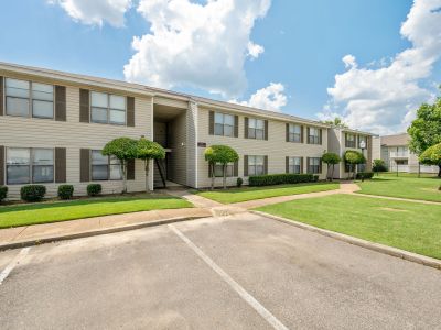 Two-story apartment building exterior featuring light vinyl siding, dark shutters, and manicured landscaping trees at The Ellington at Kirby apartments in Memphis.